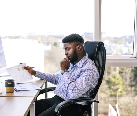 Man looks at printed graphs in front of computer screen beside window