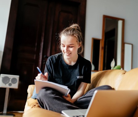 Woman taking notes in a notebook while looking at a laptop, while sitting on a couch