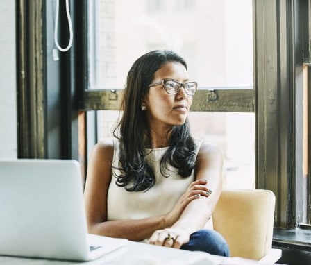 Woman looking out window, sitting at a desk with an open laptop