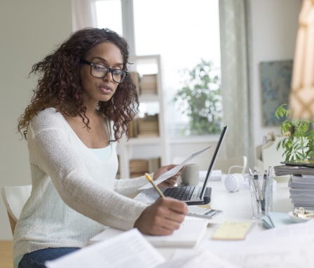 Woman writing in notebook, looking at laptop and calculator