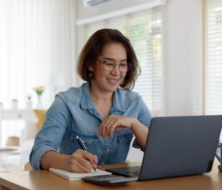 Woman smiling at laptop, writing in notebook beside calculator