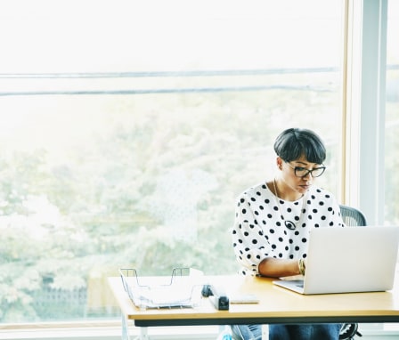 Woman working in office on laptop in front of big window