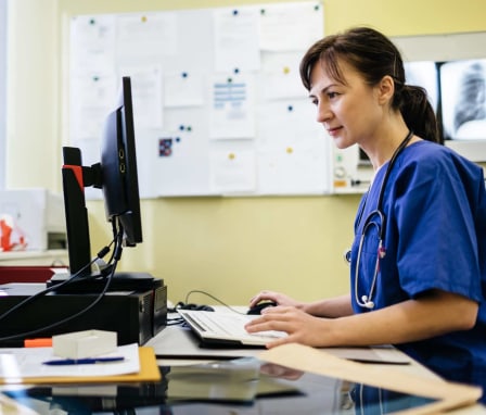 Female medical coder typing on a computer in a doctor's office. She is sitting at a desk and referencing patient medical files.