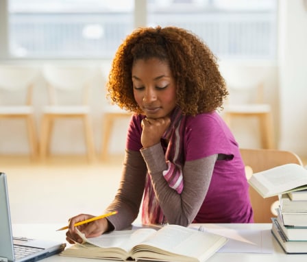 A female African-American college student sitting in a cafe and studying. She has her laptop open and is reading through textbooks.