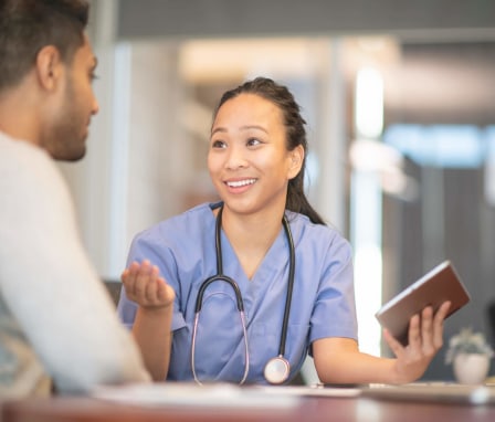 A female Asian-American psychiatric nurse holding a tablet computer is talking to her male patient. They are sitting in the doctor's office.