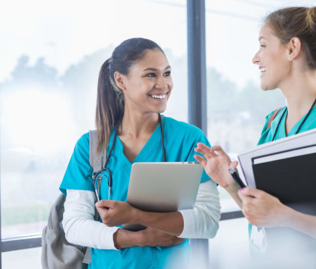 A pair of female nursing students are cheerfully chatting while walking to class together. The student pictured on the left is a young mixed-race woman with straight dark brown hair pulled back into a ponytail. She is wearing bright blue scrubs, a gray backpack, and a stethoscope around her neck, and is carrying a gray laptop in her left arm. Her classmate pictured on the right in the photo is a young Caucasian woman. She is wearing teal green scrubs and a brown backpack. She is carrying a notepad and a textbook in her left arm and a pen in her left hand. Together they are walking in a hallway lined with floor-to-ceiling glass windows with a view overlooking the school campus.