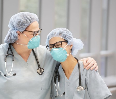 Two female, health care providers laugh while wearing light green medical scrubs, stethoscopes and N95 masks in an indoor hallway.