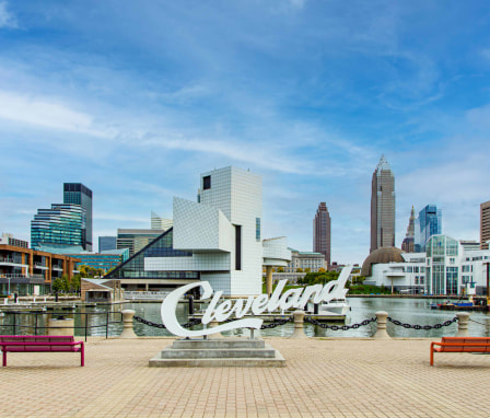 Voinovich Bicentennial Park, along the shore of Cleveland Harbor, provides a stunning view of Cleveland's beautiful skyline. The colorful buildings of Cleveland stand beneath a blue sky with wispy clouds.