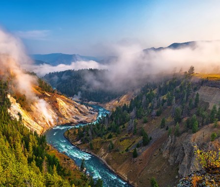 A wide angle summer morning view captured from the Calcite Springs View Area in Yellowstone National Park, Wyoming. Far below is the winding Yellowstone River, with the steaming Calcite Springs on the left bank of the river. Tall, tree-lined cliffs rise steeply on either side of the river and into the misty morning.