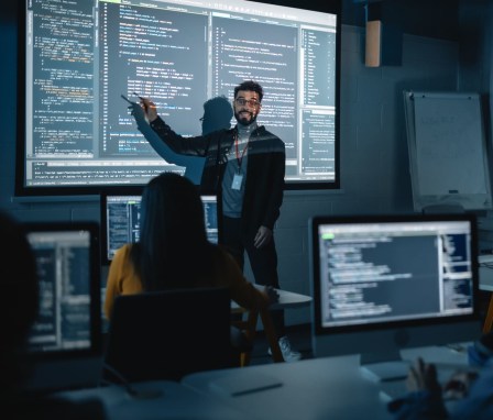 Teacher Giving Computer Science Lecture to Diverse Multiethnic Group of Female and Male Students in Dark College Room. Projecting Slideshow with Programming Code. Explaining Information Technology.