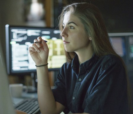 A young woman is seated at a desk surrounded by monitors displaying data, she is contemplating in this dark, moody office.