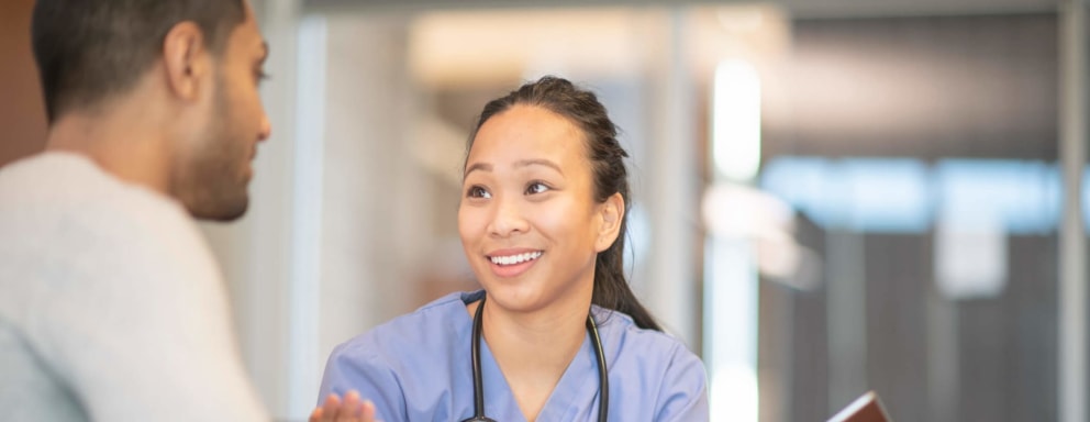 A female Asian-American psychiatric nurse holding a tablet computer is talking to her male patient. They are sitting in the doctor's office.