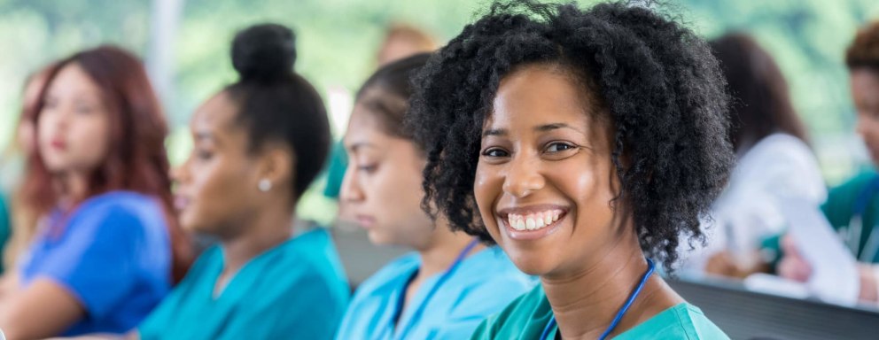 A smiling mid-adult African American nursing student is sitting in class with her colleagues. She has short, curly black hair, and is wearing teal green scrubs. Her laptop and notepad are open so she is ready to take notes during class. She is looking directly at the camera.