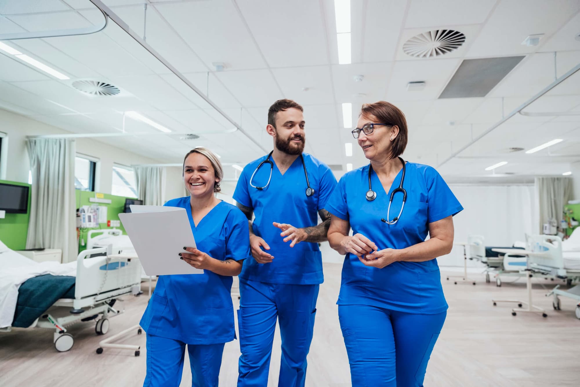 Three nurses, two female and one male, are walking and talking together through a hospital ward.