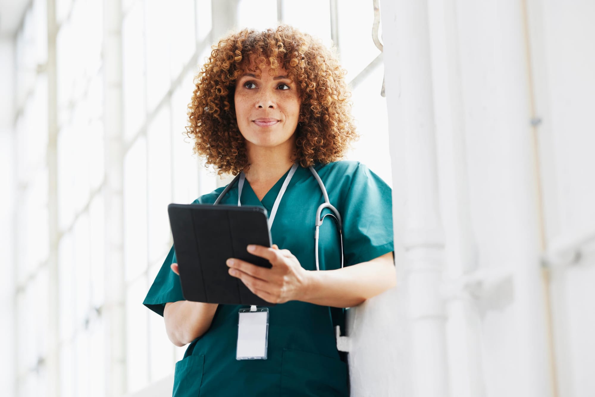 Black female nurse practitioner stands in a hospital hallway looking at patient's report on a digital tablet.