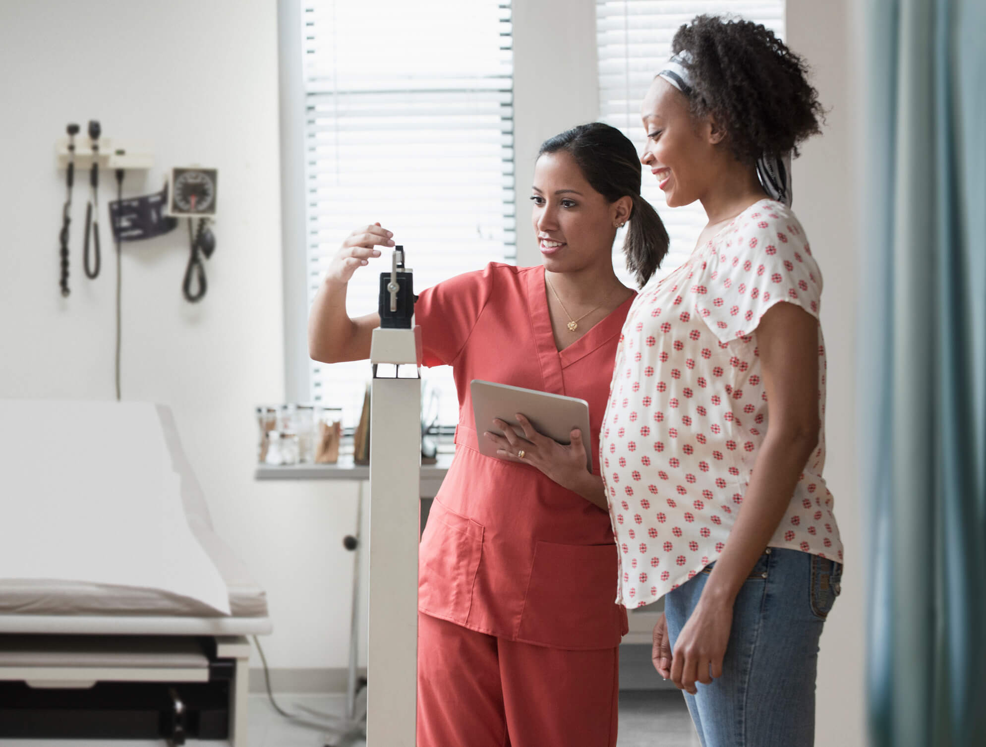 A Black female labor and delivery nurse weighs a Black pregnant patient in a hospital room.