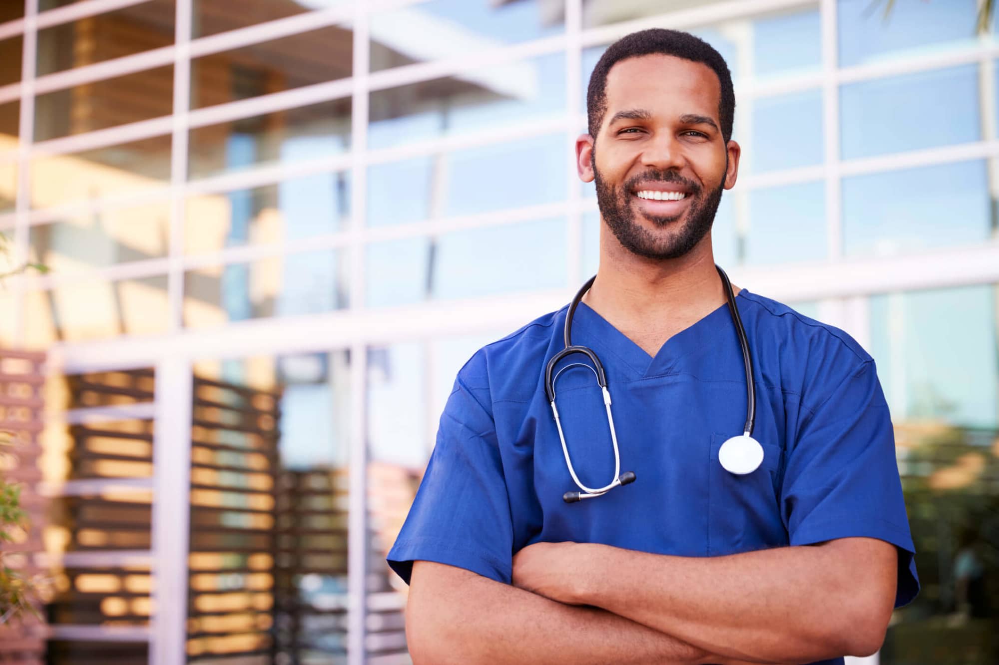 A young black male nurse standing outside a hospital smiling at the camera.