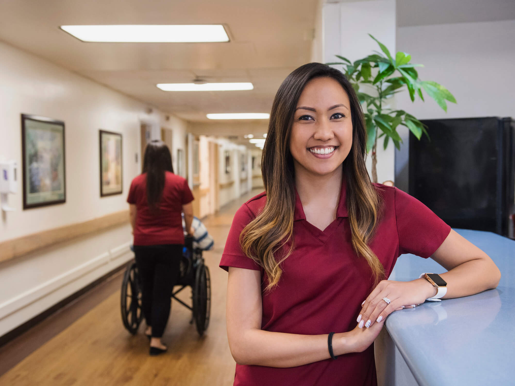 Asian-American female medical assistant smiles while standing in front of a hospital reception desk.