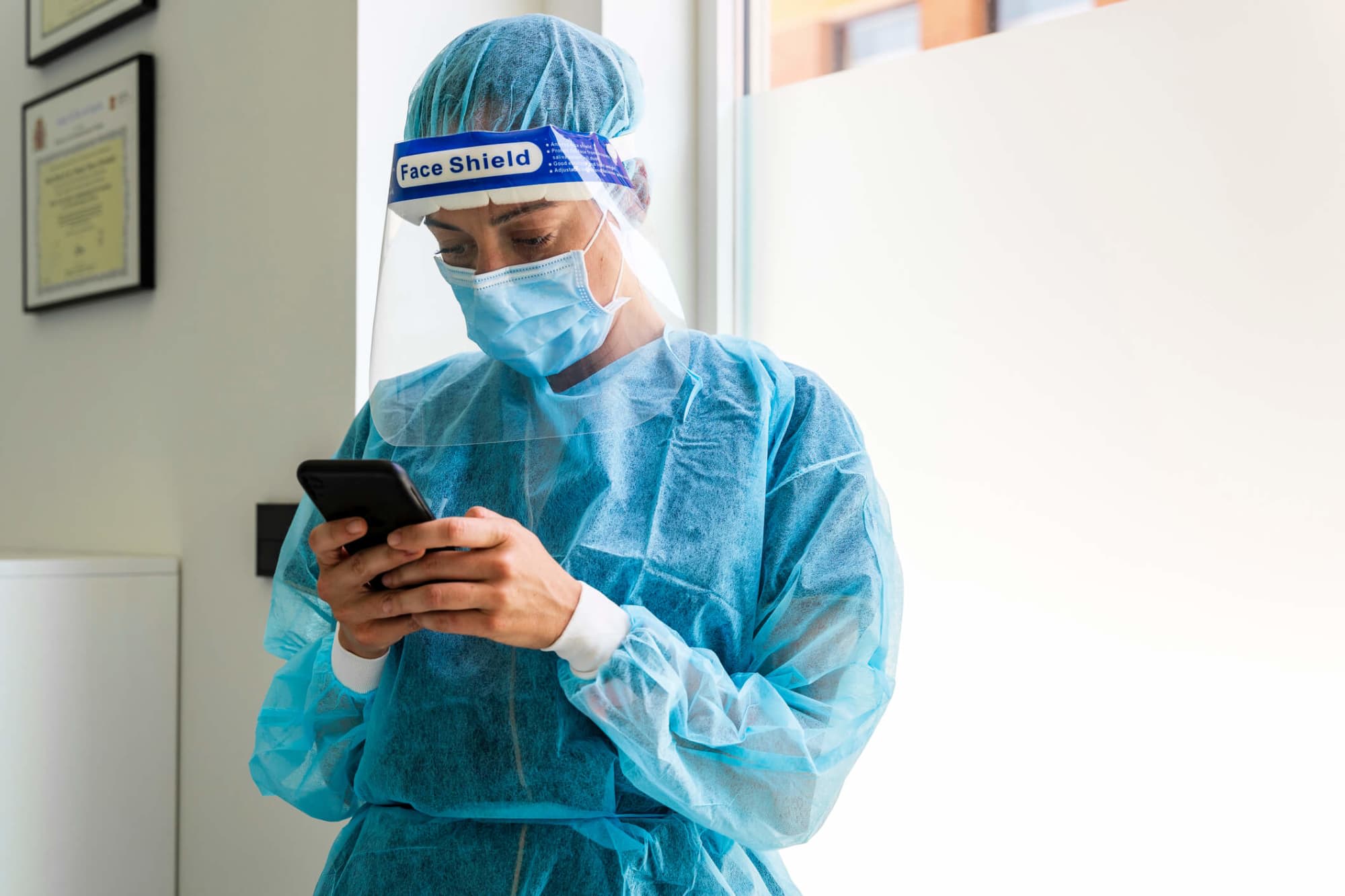Female nurse dressed in COVID-19 protective gear texting on her smart phone in a hospital.