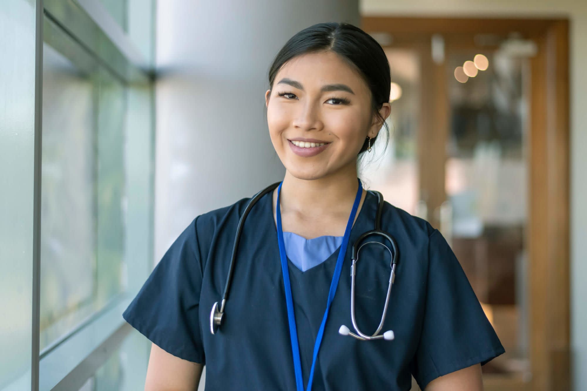 An Asian-American female nurse wearing scrubs and a stethoscope poses next to a window in a hospital corridor and smiles at the camera.