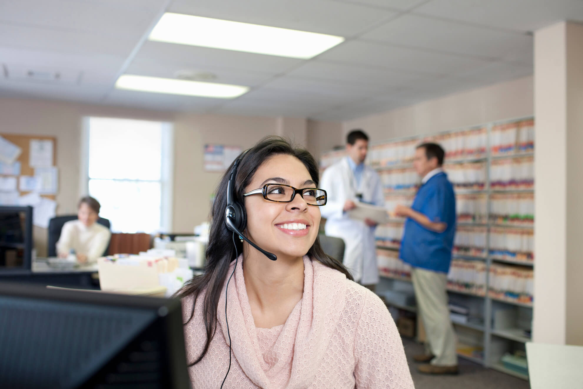 A young female medical receptionist sitting at her desk while talking with a client. Two doctors are standing while chatting in the background.