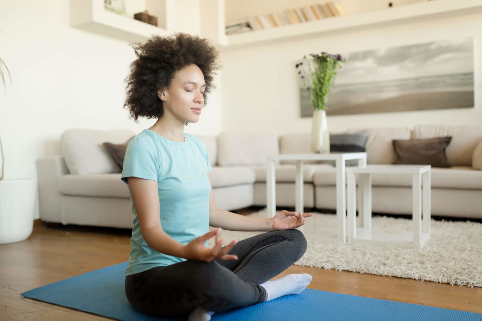 A young African American woman meditating in cross-legged position on her exercise mat in her home.