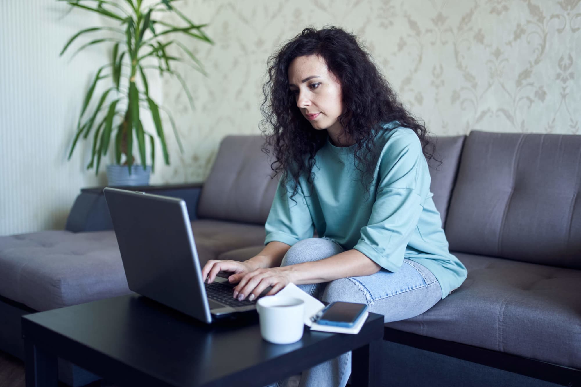 A mid-adult woman sitting at home on the couch typing on her laptop.