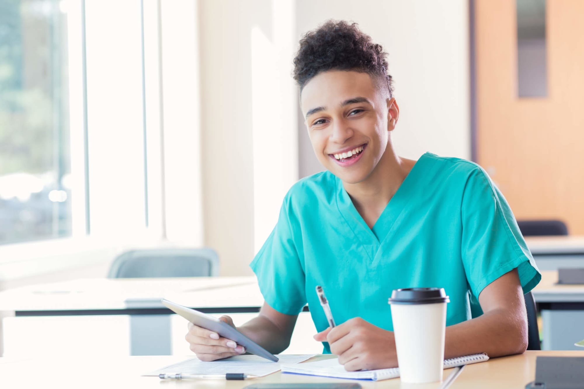 Black male nursing student sitting at a classroom desk writing notes while preparing for class. He is wearing scrubs and referencing materials from a digital tablet.