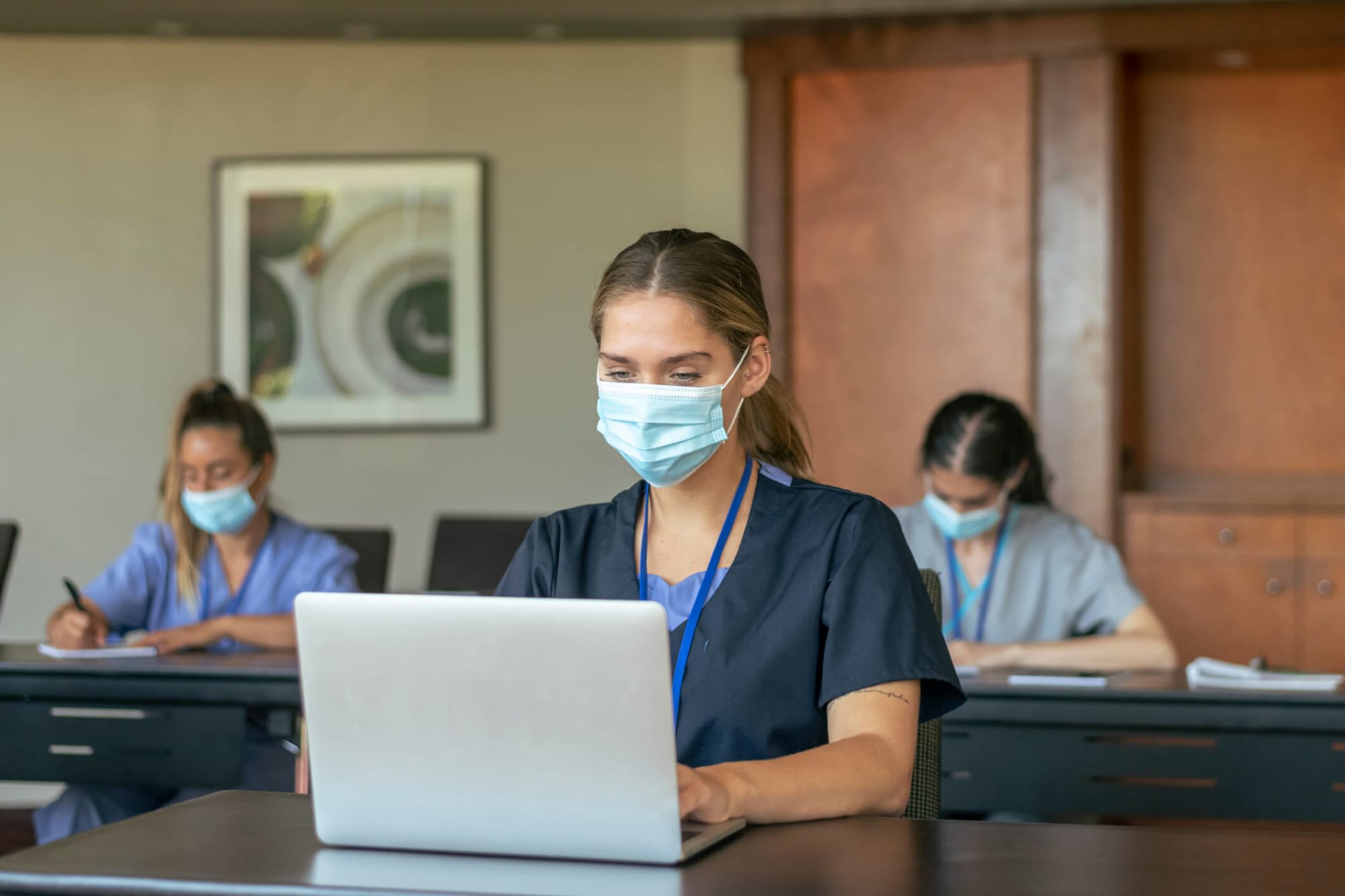 Group of nursing students wearing protective face masks back at school take notes while attending class lecture. Each individual is seated at their own desk due to Covid-19 social distancing regulations.