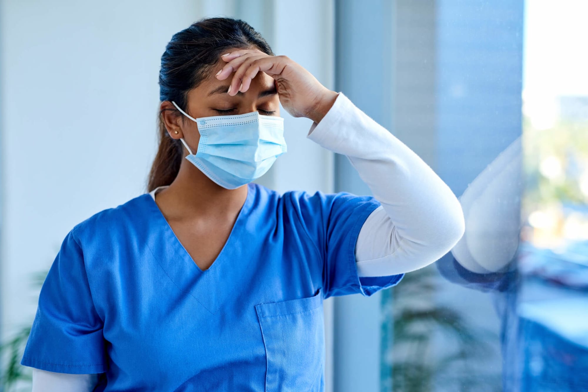 Young Asian-American female nurse with her eyes closed rubs her forehead while standing by a hospital window.