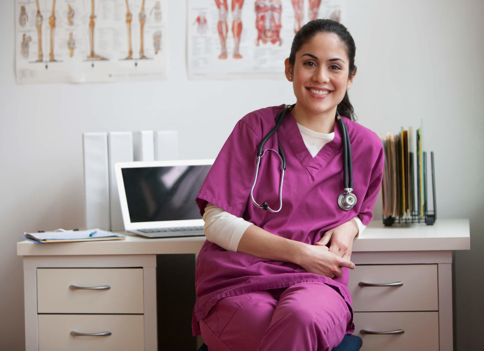 A female Hispanic nurse sitting at her desk in a clinical office. She is facing towards the camera and smiling. Her laptop and case files are on the desk behind her.