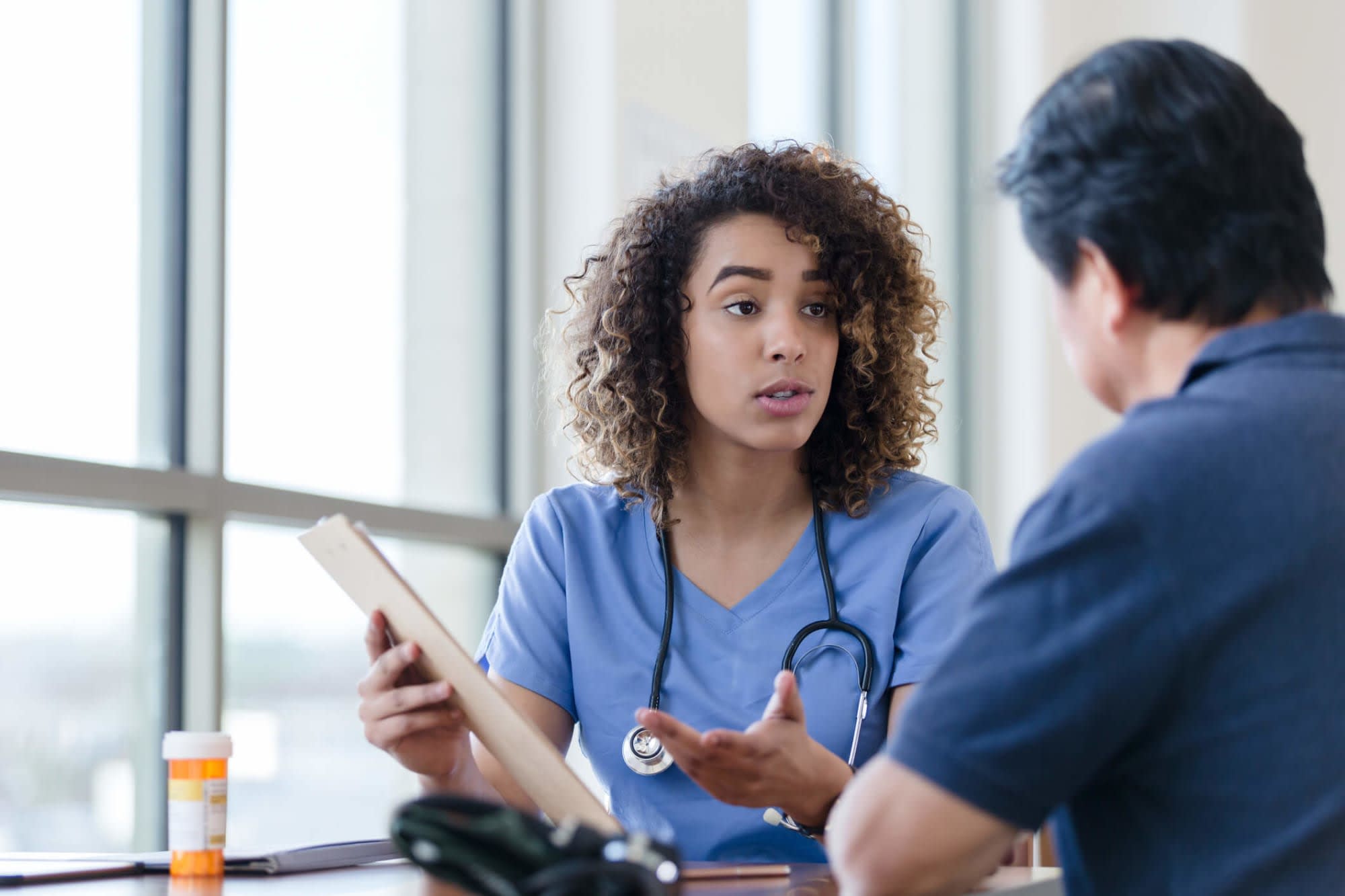 A Black female nurse case manager is discussing a care plan with a male patient. They are sitting at a desk in a hospital office. The nurse is showing the patient an outline of the plan on a clipboard.