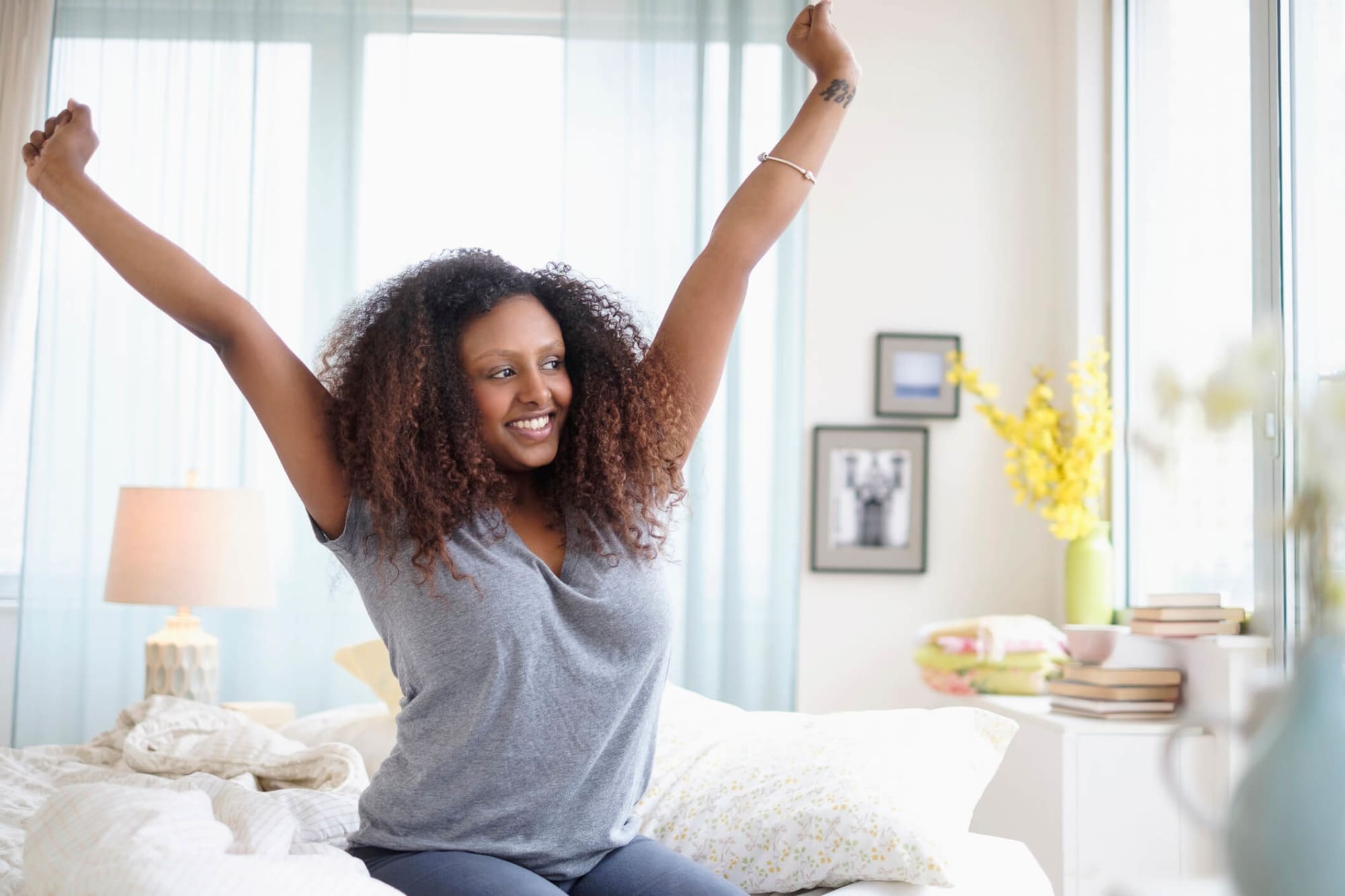 A young adult Black woman is pictured stretching on her bed on a bright sunny day. She is smiling and looks as if she has just woken up from a restful sleep.