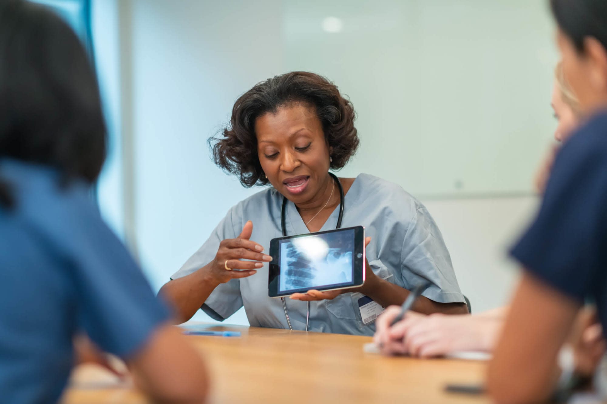 A mature Black female nurse researcher is leading a meeting with other nurses. She is showing the group a medical x-ray on a digital tablet. The multi-ethnic group of medical professionals is seated around a table in a conference room.