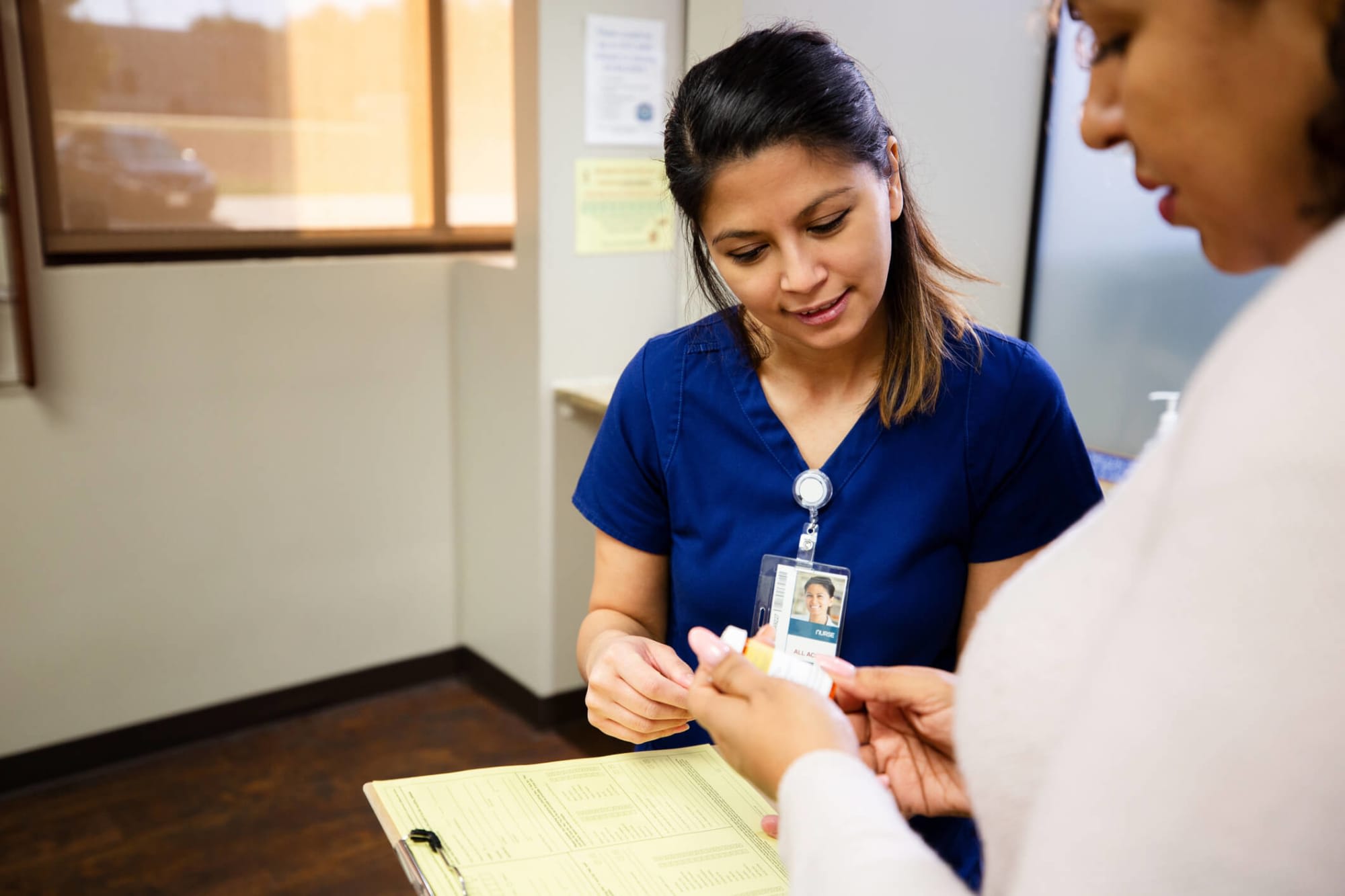 A mid-adult Hispanic female nurse is chatting with a physician behind the administrative desk of the clinic. She is holding a patient file in her left hand and consulting with the physician about the patient's medication.