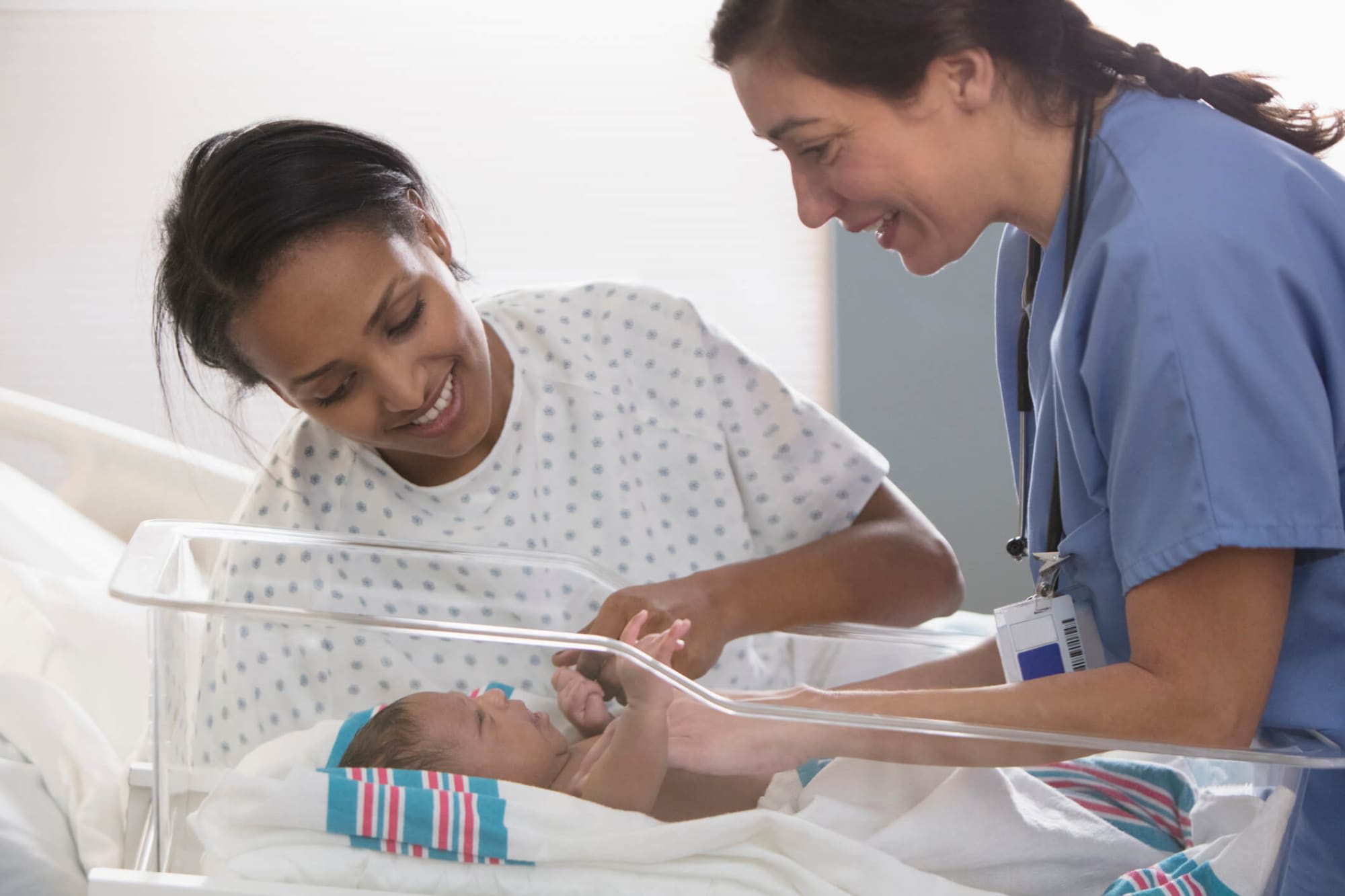 A mid-adult female nurse has just carted a newborn child to its mother's hospital bedside. The mother is a mid-adult Black woman wearing a hospital gown. She is smiling and leaning over her bedside to greet her newborn child.