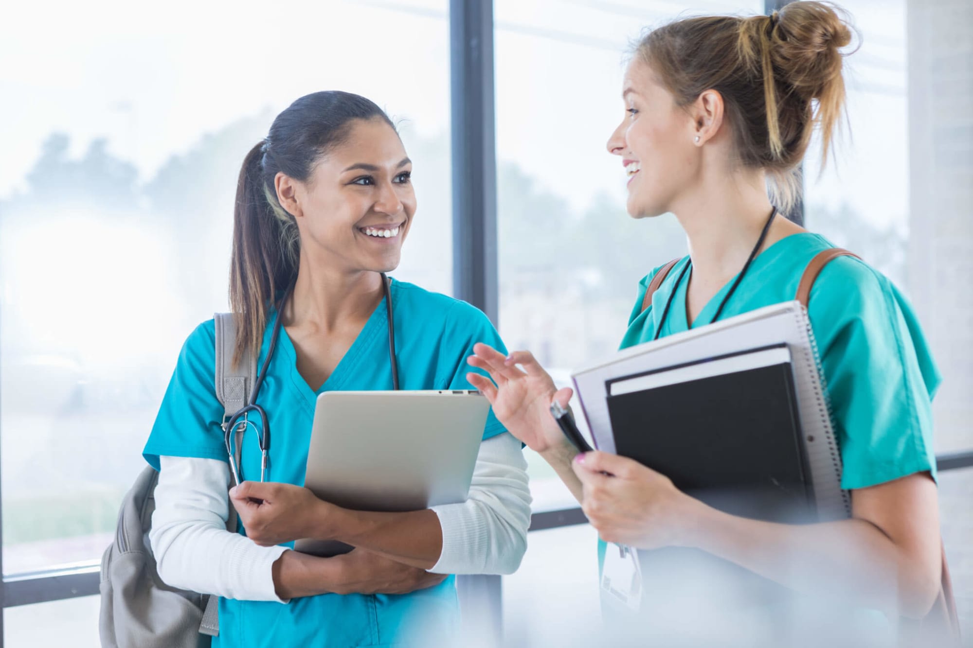 A pair of female nursing students are cheerfully chatting while walking to class together. The student pictured on the left is a young mixed-race woman with straight dark brown hair pulled back into a ponytail. She is wearing bright blue scrubs, a gray backpack, and a stethoscope around her neck, and is carrying a gray laptop in her left arm. Her classmate pictured on the right in the photo is a young Caucasian woman. She is wearing teal green scrubs and a brown backpack. She is carrying a notepad and a textbook in her left arm and a pen in her left hand. Together they are walking in a hallway lined with floor-to-ceiling glass windows with a view overlooking the school campus.