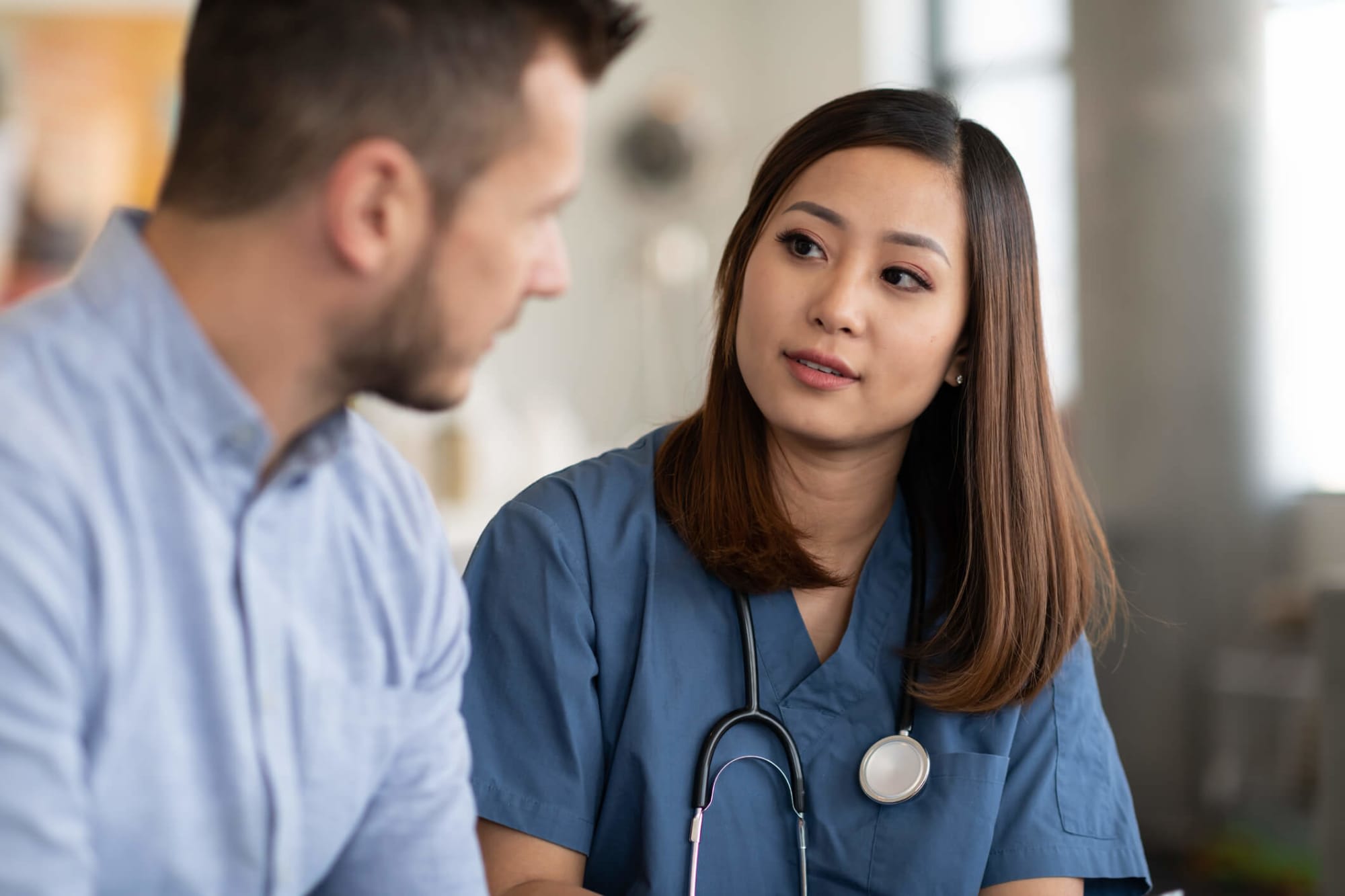 A young female Asian-American nurse and a young male Caucasian patient are chatting inside a clinical office. The nurse is listening to the man while holding a tablet computer she uses to take notes. She has long brown hair and is wearing blue scrubs and a stethoscope.