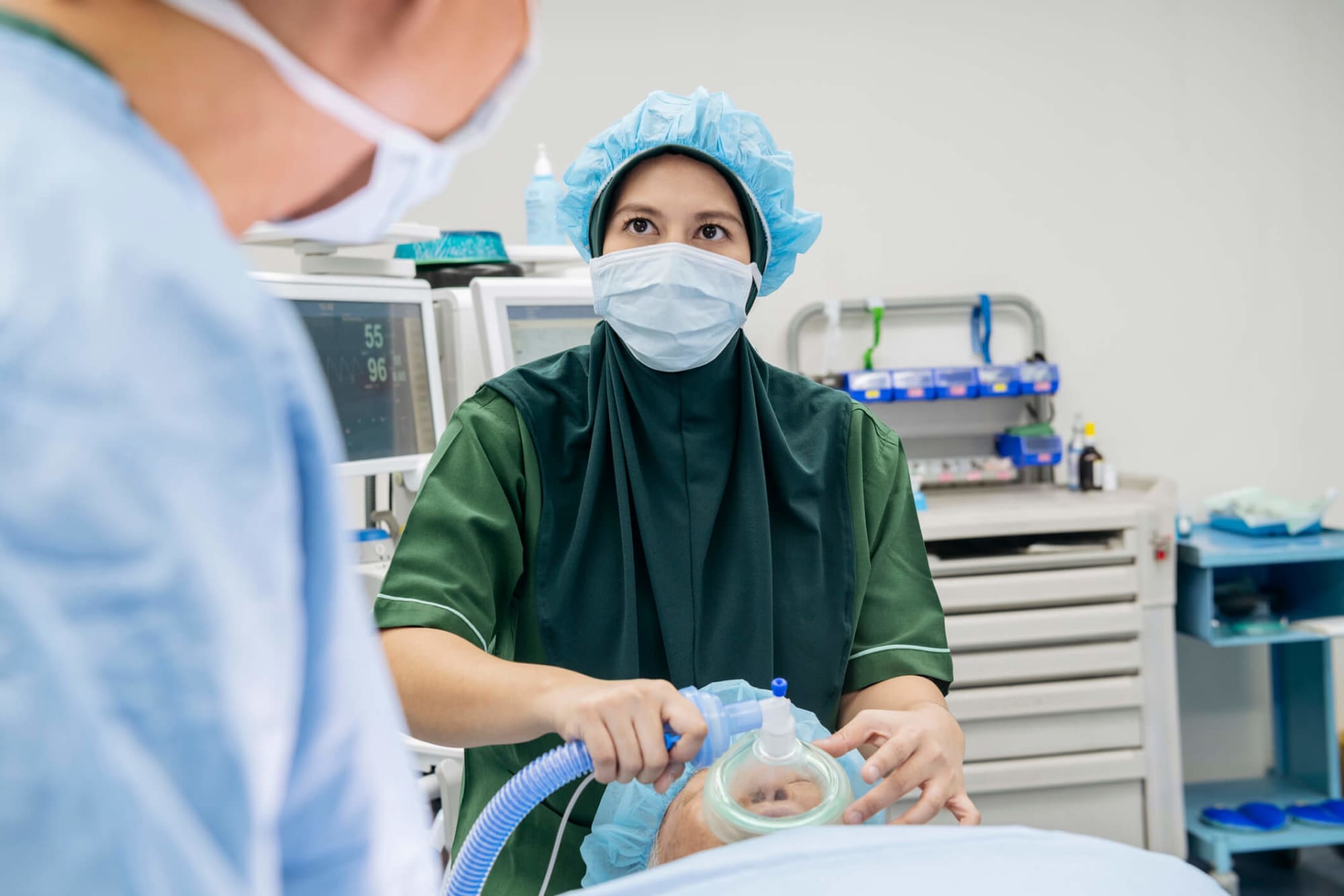 A female nurse anesthetist places an oxygen mask on a senior patient while sedating him in preparation for a surgical procedure in a hospital operating room. The young Malaysian nurse is wearing a dark green hijab over her green scrubs, and has a mask and medical cap on. A heart monitor and medical equipment are in the background. The nurse is looking towards the doctor in the foreground while awaiting his instructions.