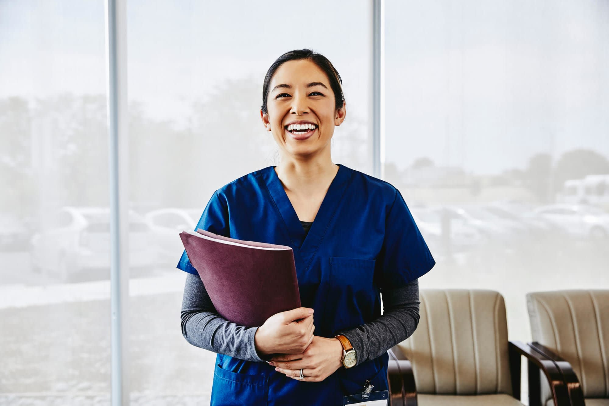 Picture of a smiling Asian-American nurse standing in a hospital waiting room. She has black hair pulled back into a ponytail, and is wearing dark blue scrubs. She is carrying a patient file in her right arm.