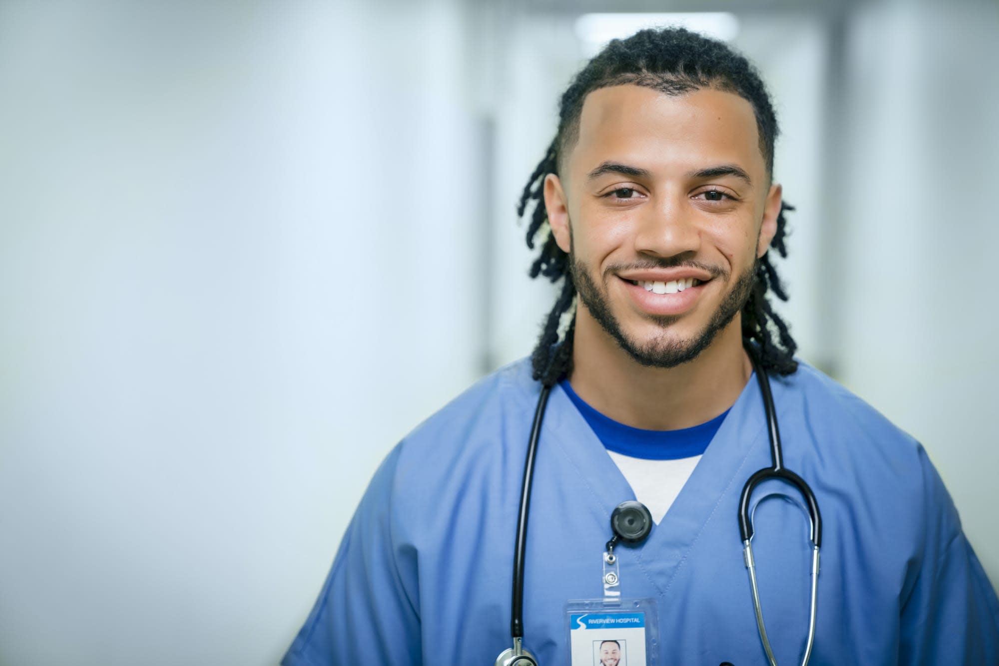 A young mixed race nurse is standing in a hospital hallway. He is wearing light blue scrubs and is looking at the camera and smiling.