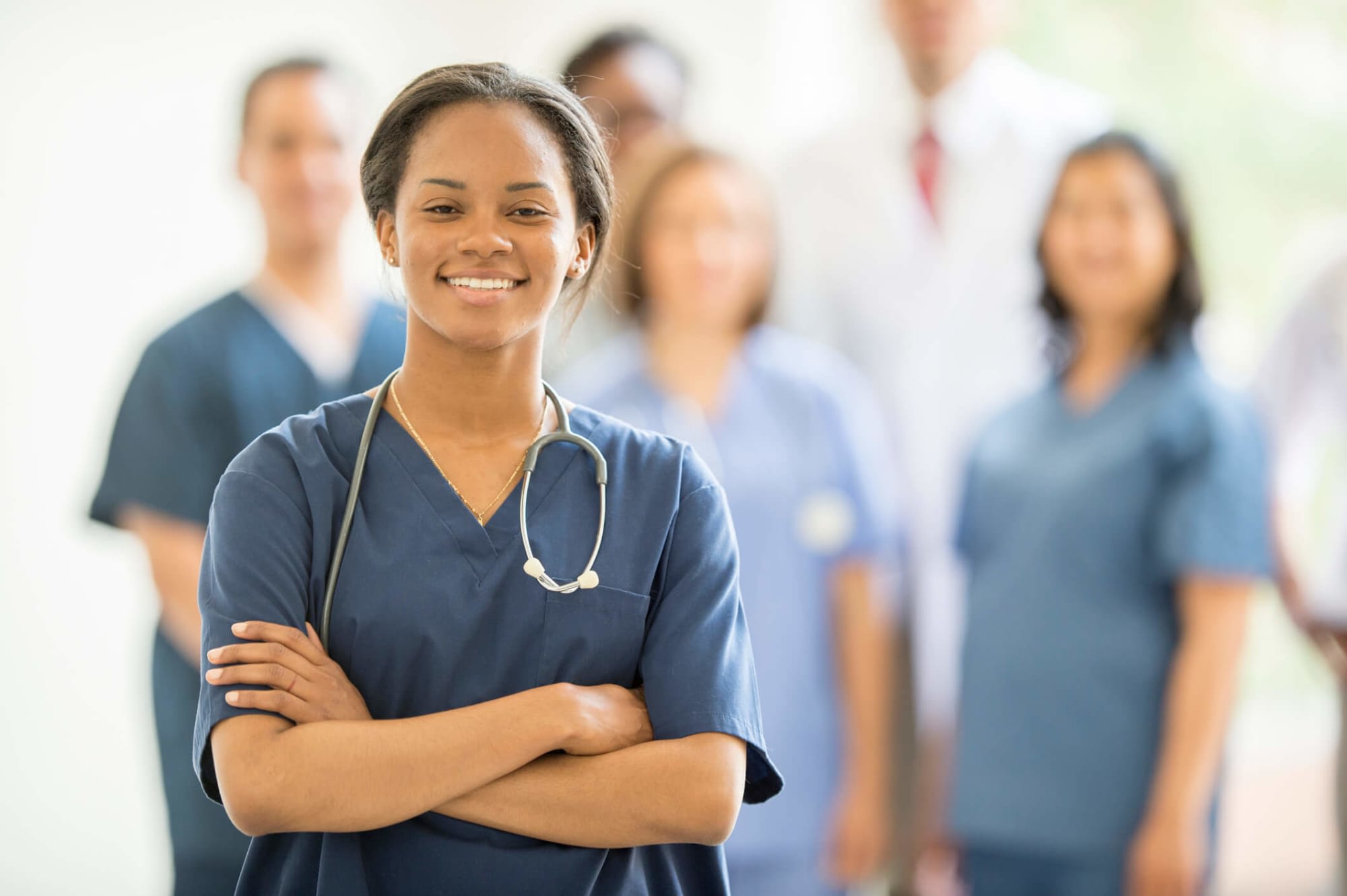 A young Black female nurse is smiling and looking at the camera. She has long brown hair pulled back into a ponytail, and is wearing dark bluish-gray scrubs. Behind her, a multi-ethnic group of doctors and nurses are standing together inside of the hospital.