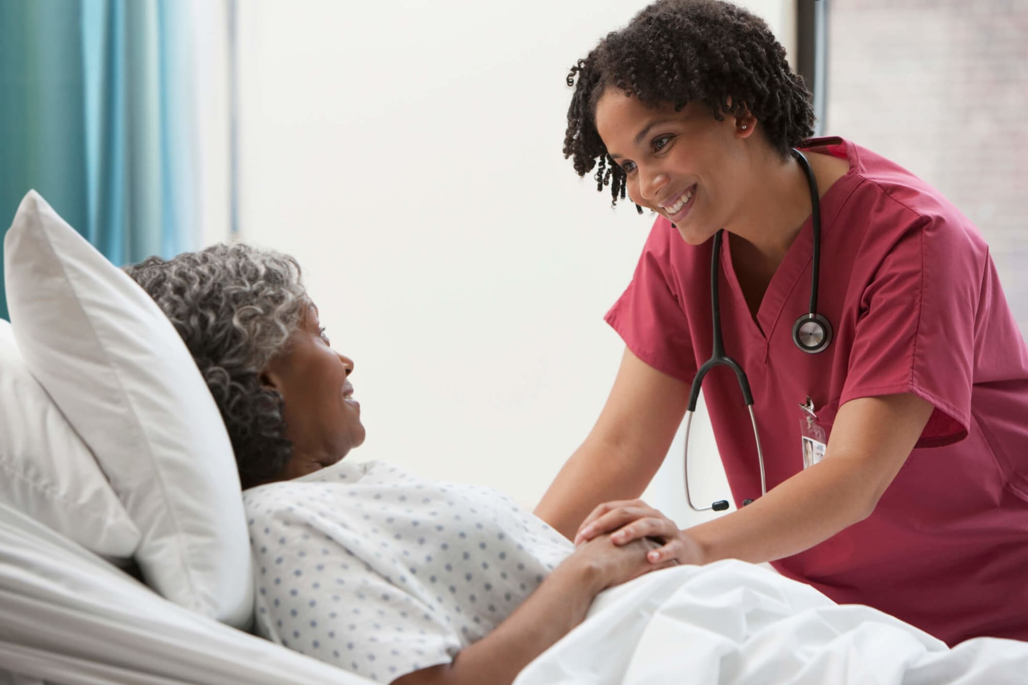 A young Black female nurse is checking in on one of her elderly patients during her hospital rounds. She has short, curly black hair and is wearing pinkish-red colored scrubs. The patient is an elderly black female wearing a hospital gown. The nurse is standing beside the patient's bedside, smiling and chatting with her.