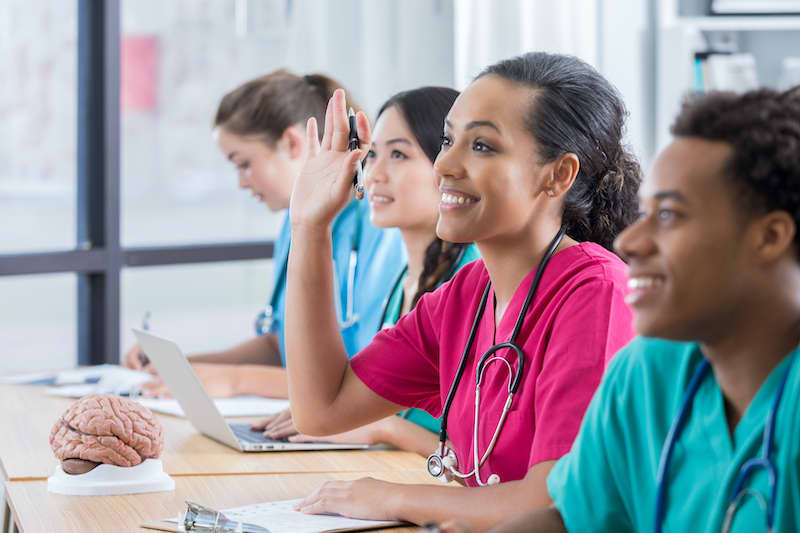 Smiling young female African American student raises her hand to ask a question in class. She is sitting in the middle of three other students at a string of desks. Two other students are looking forward and smiling and another is writing at her desk.