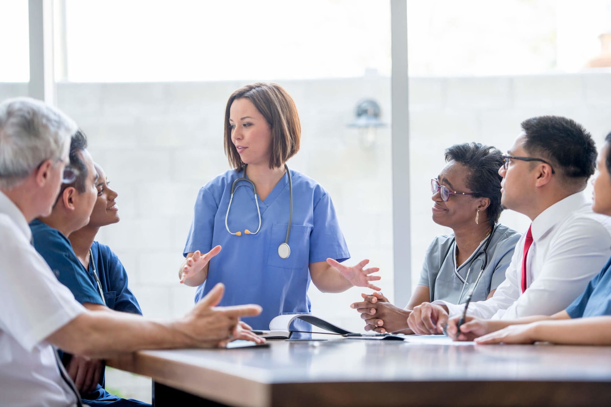 A multi-ethnic group of medical professionals are sitting together at a table discussing a public health case. A young white female nurse is leading the conversation. She has short brown hair and is wearing her light blue scrubs.