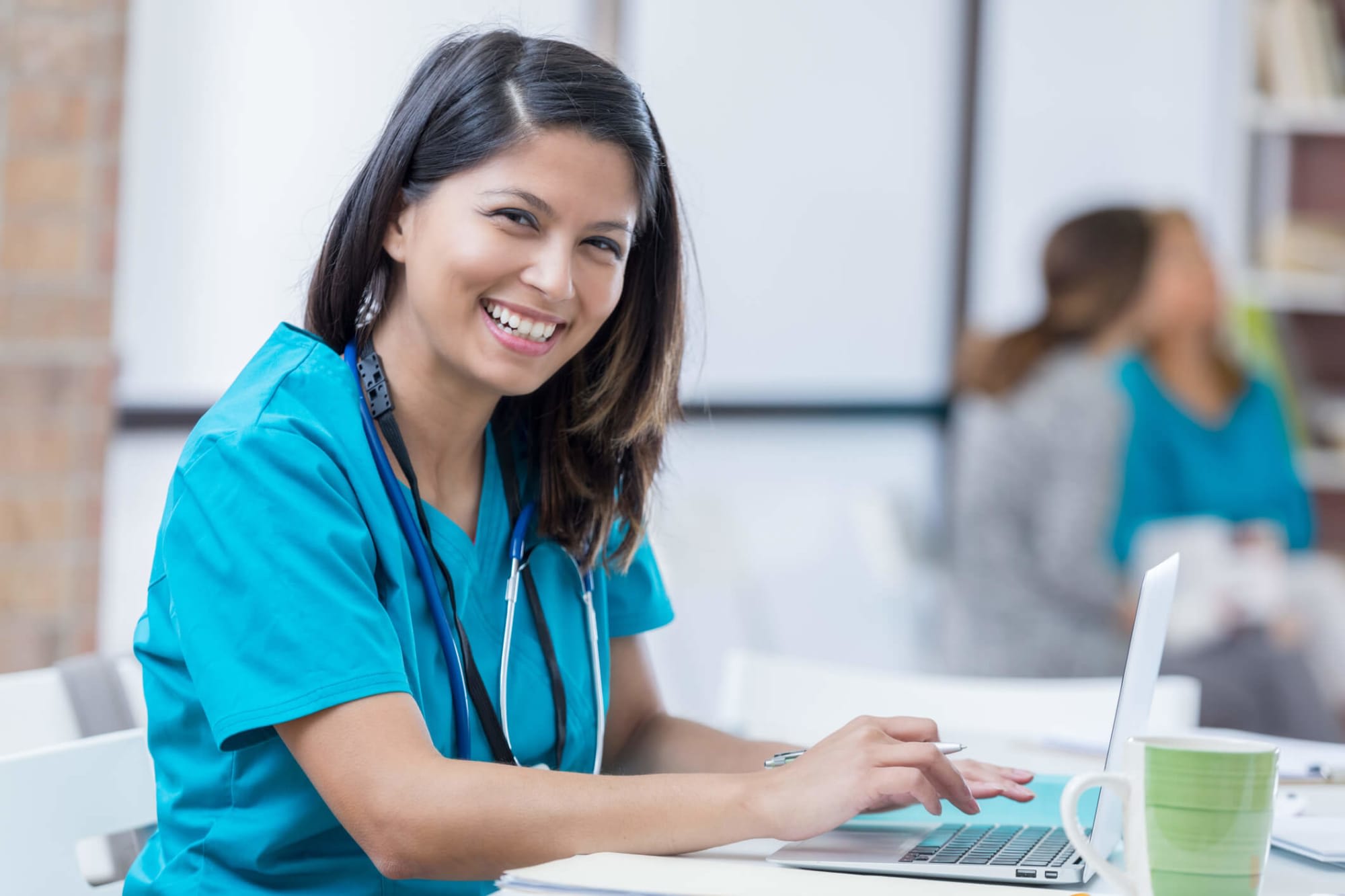 A female Filipino nurse is sitting down at her laptop to study. She is wearing scrubs and a stethoscope. She is smiling at the camera. Image credit: SDI Productions / E+ / Getty Images