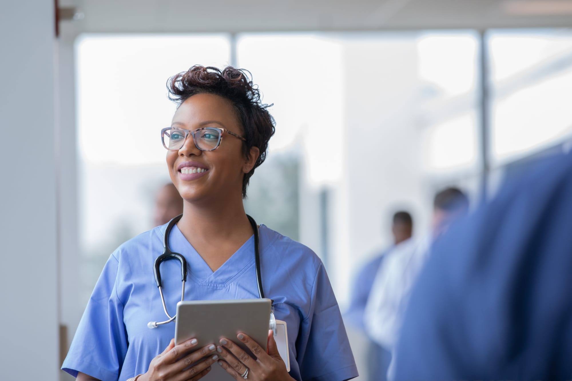 Female nurse smiles while staring out window in hospital hallway and holding digital tablet with electronic patient file