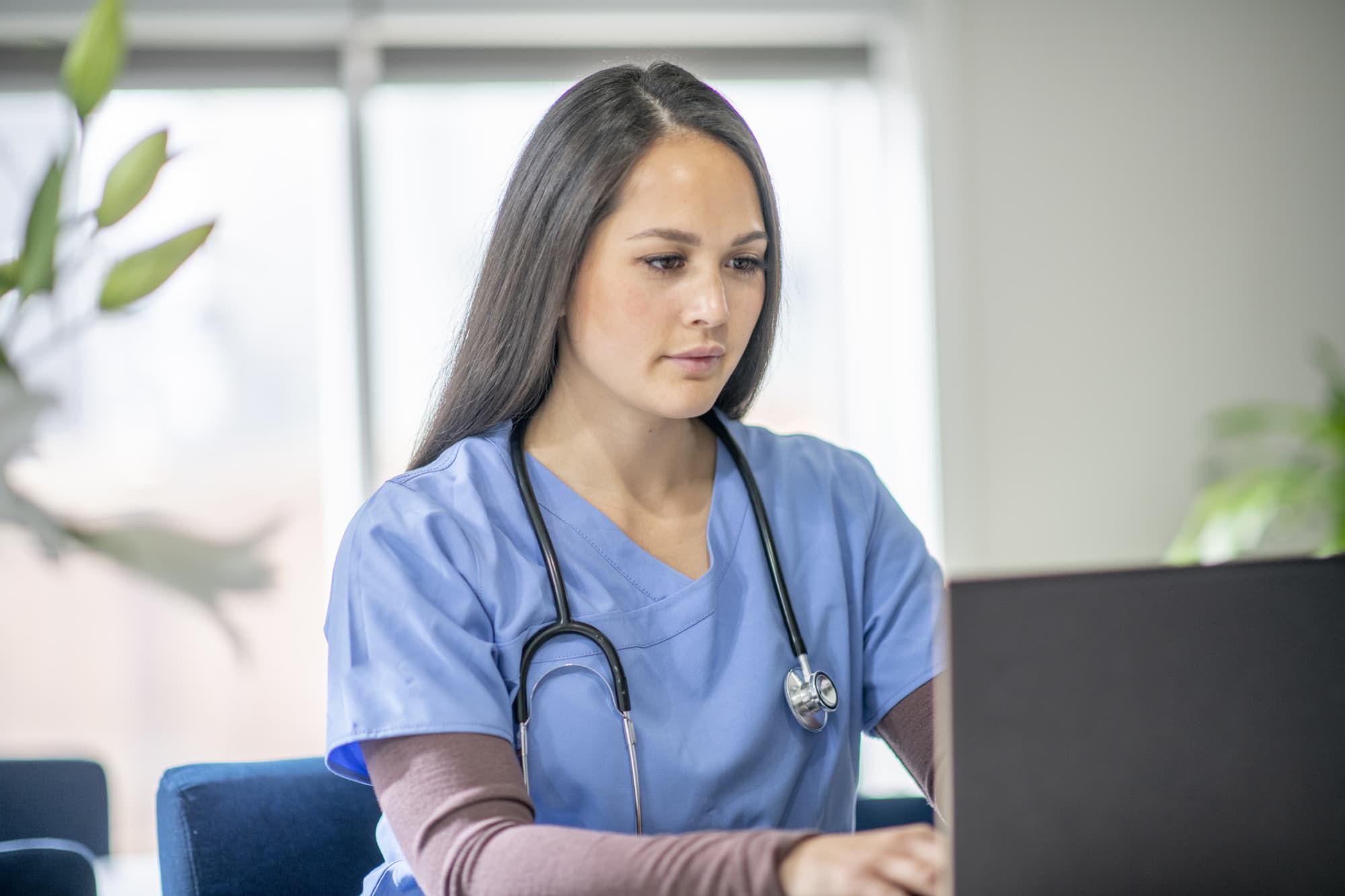 A nurse wearing scrubs interacts with a patient through a video call on her computer.