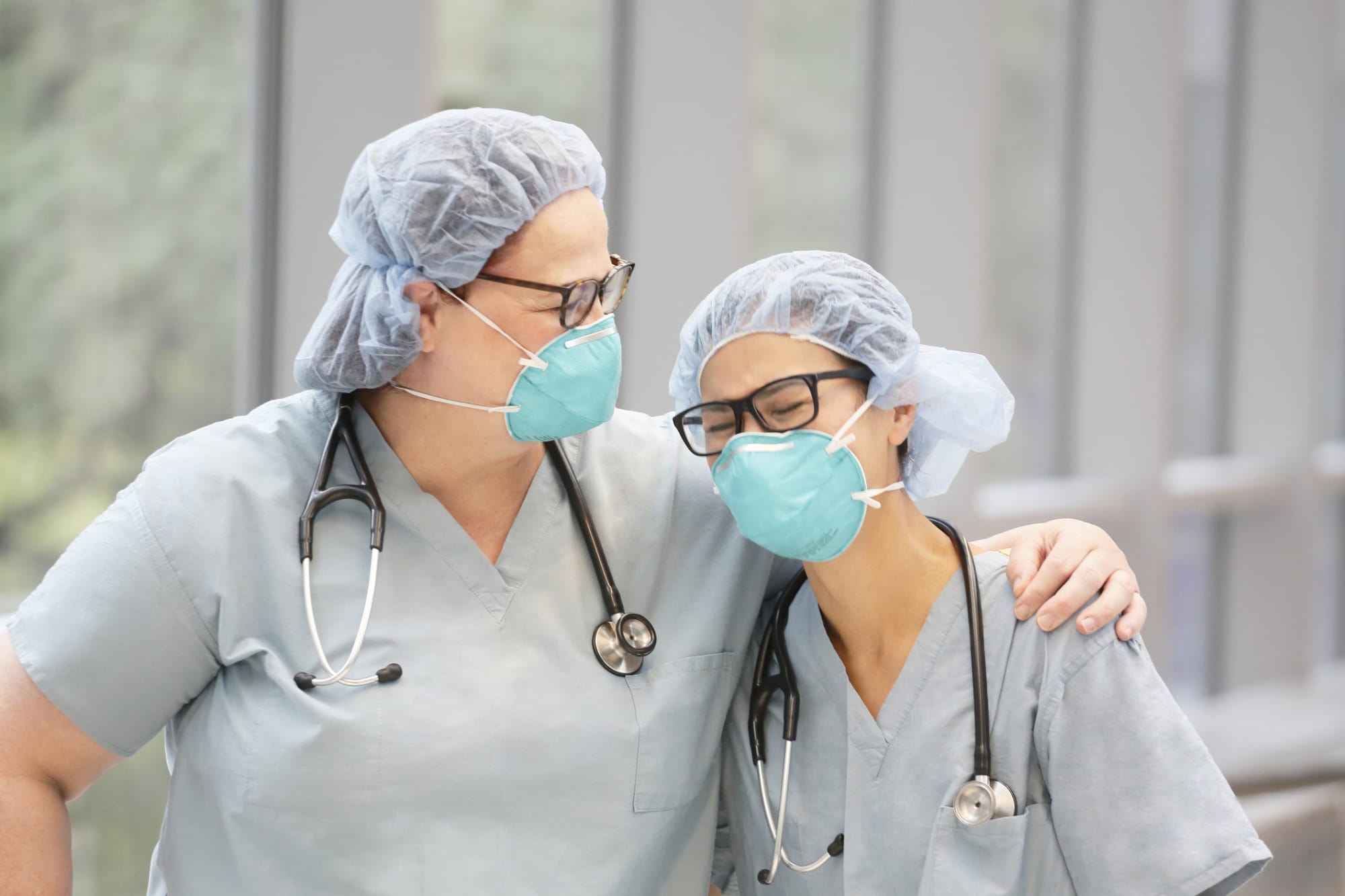 Two female, health care providers laugh while wearing light green medical scrubs, stethoscopes and N95 masks in an indoor hallway.