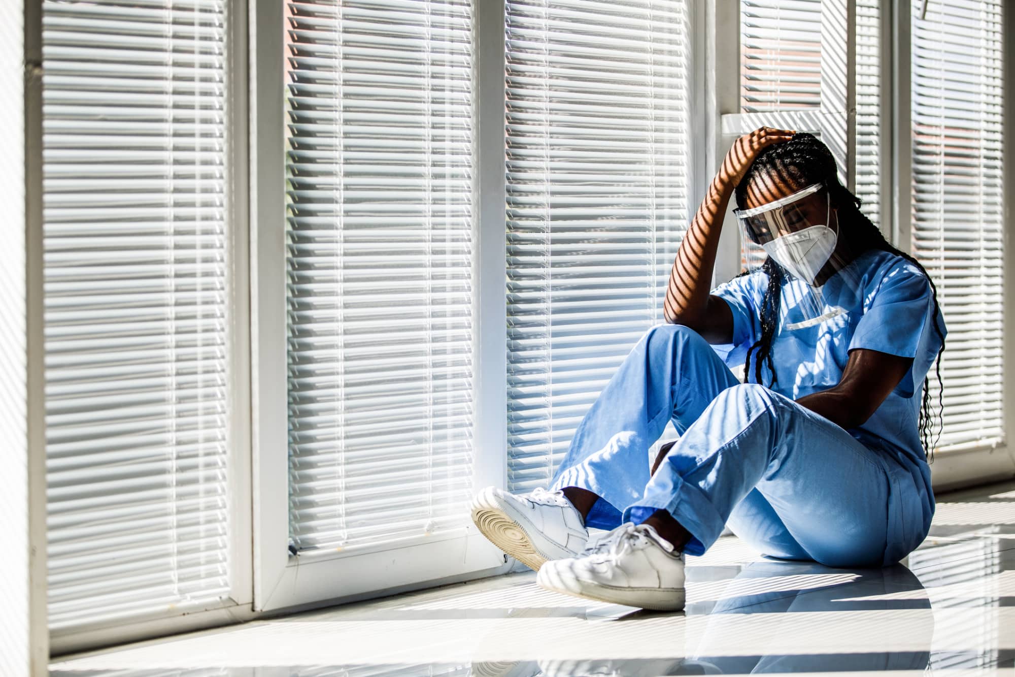Overwhelmed nurse wearing PPE sitting down in a hospital hallway